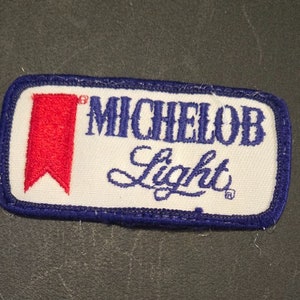 May include: A white embroidered patch with blue stitching around the edges. The patch features a red banner and the text "Michelob Light" in blue cursive lettering.