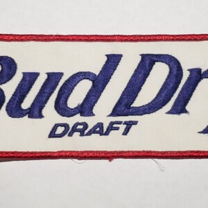 May include: A white and red embroidered patch with the text "Bud Dry Draft" in blue lettering and a registered trademark symbol.