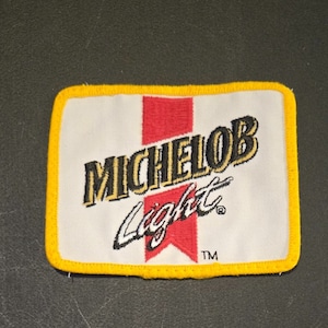 May include: A white embroidered patch with a yellow border. The patch features the text "Michelob Light" in black and gold lettering with a red stripe behind the text.