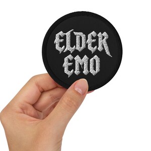 Pode incluir: Um patch redondo preto com o texto bordado "ELDER EMO" em letras góticas brancas. O patch é segurado em uma mão, mostrando o design e a textura das letras bordadas.