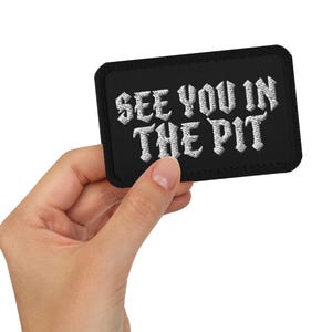 May include: Black rectangular patch with the text "SEE YOU IN THE PIT" in white, gothic-style lettering. The patch is held in a hand.