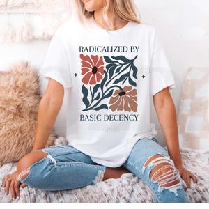 May include: A white short-sleeve t-shirt featuring a graphic print with the text RADICALIZED BY BASIC DECENCY. The design includes two stylised flowers in terracotta and muted brown, surrounded by dark teal leafy foliage on a white background.