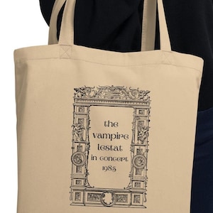 May include: A beige tote bag featuring a black and white illustration of an ornate archway. The text "the vampire lestat in concert 1985" is printed within the archway.