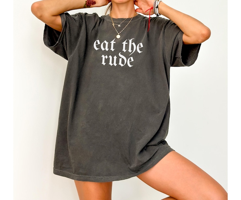 Op de afbeelding: Donkergrijs t-shirt met de tekst "eat the rude" in een gotisch lettertype. Het shirt heeft korte mouwen en een relaxte pasvorm. De stof lijkt zacht en licht vervaagd, wat het een vintage uitstraling geeft.