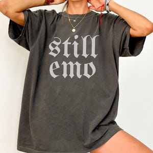 以下が含まれることがあります： ゴシック体で「still emo」と白でプリントされたダークグレーのオーバーサイズTシャツ