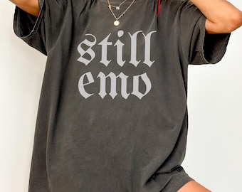 May include: A dark gray oversized t-shirt with the words "still emo" printed in white in a gothic font.