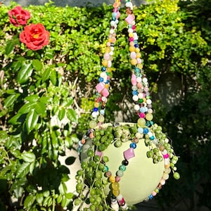Puede incluir: Un macetero colgante verde con una cadena de cuentas de varios colores y materiales. El macetero está lleno de una planta suculenta verde.