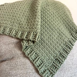 May include: A sage green crocheted blanket with a textured stitch pattern. The blanket is folded, showcasing the detailed edge and overall design. The soft, handmade blanket is likely for a baby or small child.