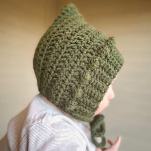 Könnte beinhalten: Handgefertigte, olivgrüne Häkelmütze mit spitzer Spitze und Ohrenklappen. Die Mütze ist aus strukturiertem Garn gefertigt und hat einen Bindverschluss unter dem Kinn. Die Mütze ist für ein Baby oder Kleinkind konzipiert.