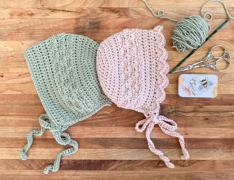 May include: Two crocheted baby bonnets, one in a light green color and the other in a light pink color, with matching ties. The bonnets are laid out on a wooden surface with a ball of yarn, a crochet hook, and a small tin box with a bee design.