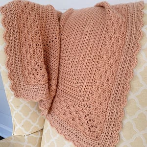 Peut inclure: Une couverture en crochet de couleur rose poussiéreux avec un bord festonné. La couverture est drapée sur une chaise avec un tissu à motifs.