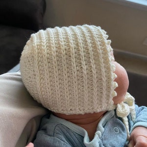 May include: A white crocheted baby bonnet with a scalloped edge and a tie. The bonnet is worn by a baby lying on a light brown surface.