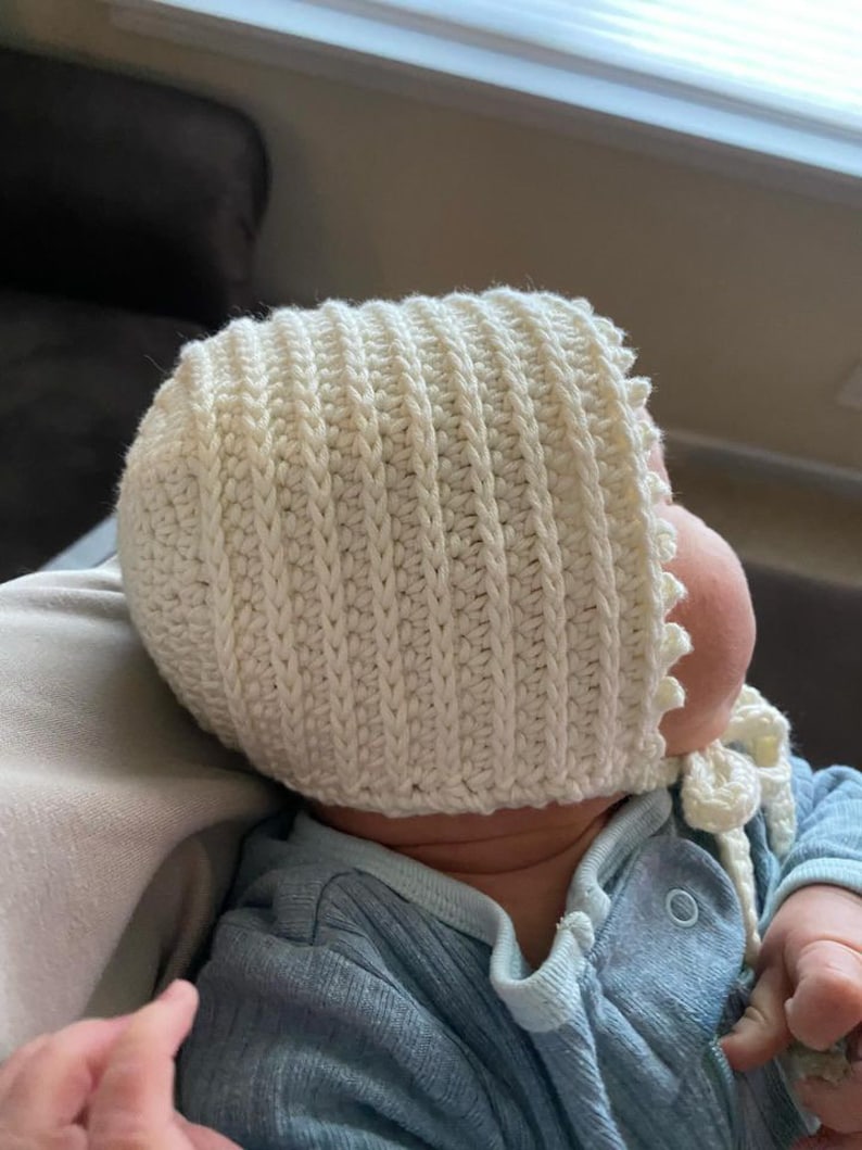 May include: A white crocheted baby bonnet with a scalloped edge and a tie. The bonnet is worn by a baby lying on a light brown surface.