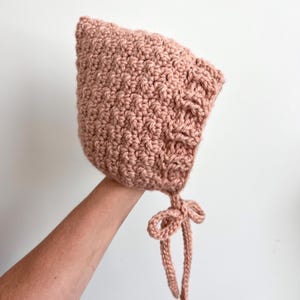 Op de afbeelding: Een handgemaakte, oudroze gehaakte muts met een textuursteekpatroon. De muts heeft een puntige kroon en delicate strikjes die onder de kin een strik vormen. Deze zachte, comfortabele hoed is voor baby's en jonge kinderen.