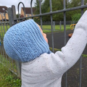 May include: A baby wearing a light blue crocheted bonnet with a white sweater. The baby is standing near a metal fence.