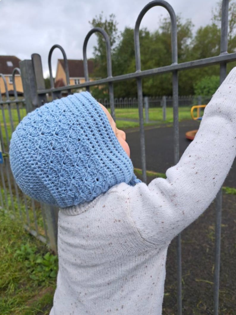 May include: A baby wearing a light blue crocheted bonnet with a white sweater. The baby is standing near a metal fence.