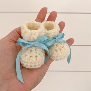 May include: A pair of crocheted baby booties in white with blue ribbon bows.