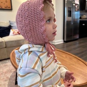 May include: A young child wearing a pink crocheted bonnet with a scalloped edge. The bonnet has a long tie that hangs down the back of the child's neck. The child is wearing a striped hoodie with a white background and brown and purple stripes.