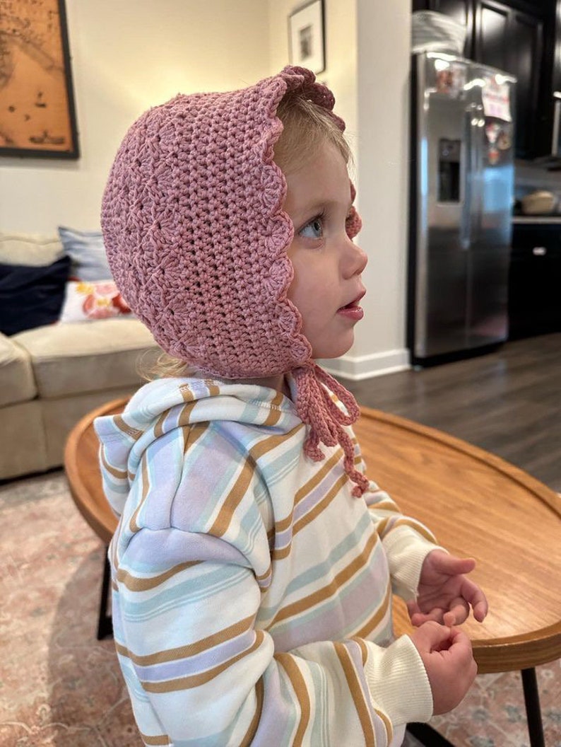May include: A young child wearing a pink crocheted bonnet with a scalloped edge. The bonnet has a long tie that hangs down the back of the child's neck. The child is wearing a striped hoodie with a white background and brown and purple stripes.