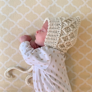 May include: A baby wearing a cream-colored crocheted hat with a unique braided design. The hat has a long tie that is tied under the chin. The baby is lying on a white and cream patterned sheet.