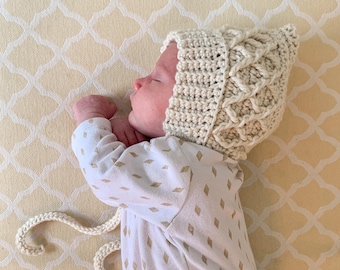 May include: A baby wearing a cream-colored crocheted hat with a unique braided design. The hat has a long tie that is tied under the chin. The baby is lying on a white and cream patterned sheet.