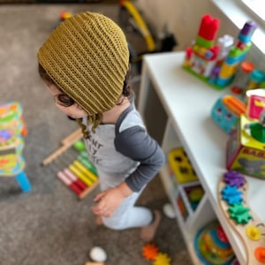 May include: A child wearing a mustard yellow crocheted bonnet with ties. The child is standing in a room with a white bookshelf and various colorful toys.