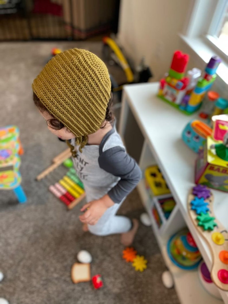May include: A child wearing a mustard yellow crocheted bonnet with ties. The child is standing in a room with a white bookshelf and various colorful toys.