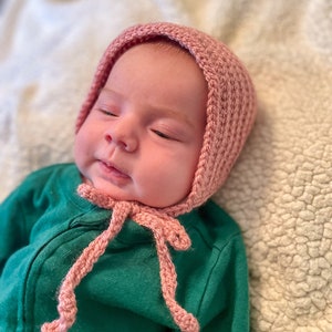 May include: A baby wearing a pink crocheted bonnet with a matching tie. The baby is lying on a white fluffy blanket.