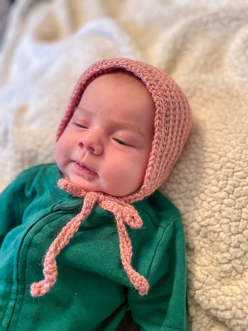 May include: A baby wearing a pink crocheted bonnet with a matching tie. The baby is lying on a white fluffy blanket.