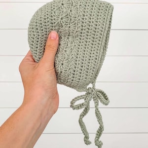 May include: A sage green crocheted baby bonnet with a tied bow at the bottom.