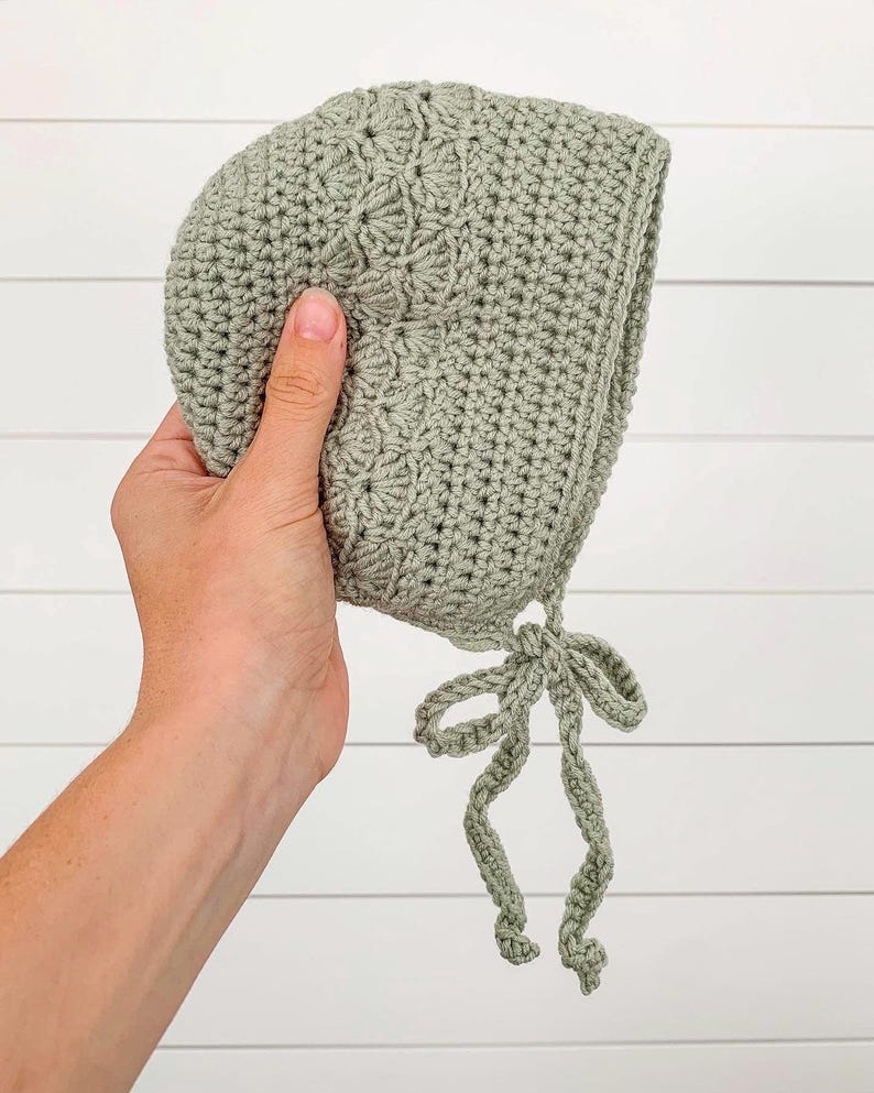 May include: A sage green crocheted baby bonnet with a tied bow at the bottom.