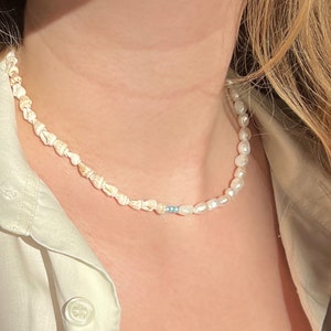 Könnte beinhalten: Eine Halskette mit einer Kombination aus Muscheln und Süßwasserperlen. Die Halskette hat einen Abschnitt mit kleinen, hellfarbigen Muscheln, gefolgt von ein paar blauen Perlen und dann einer Reihe weißer Perlen. Die Halskette wird von einer Person getragen.