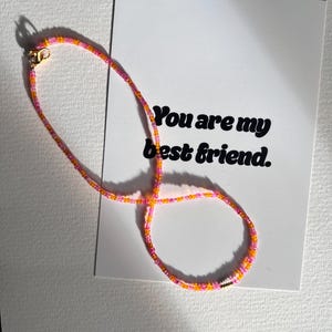 Könnte beinhalten: Eine Halskette aus rosa, orangefarbenen und gelben Perlen. Die Halskette wird auf einer weißen Karte mit dem Text "You are my best friend" gezeigt. Die Halskette hat einen goldenen Verschluss.