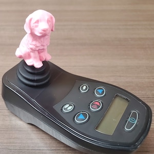 May include: A pink plastic dog figurine sits on top of a black joystick with multiple buttons and a digital display.