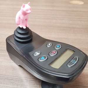 May include: A black joystick controller with a pink cat figurine on top. The controller has several buttons and a small display screen. The cat is sitting upright, facing forward. The controller is on a wooden surface.