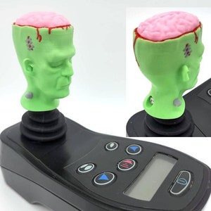 May include: A green Frankenstein-themed joystick topper with a pink brain and red blood drips. The topper is mounted on a black controller with buttons and a small screen. A novelty item with a unique design.