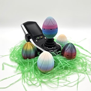 May include: A black joystick with decorative, egg-shaped toppers. Several multi-coloured Easter eggs are arranged on green paper grass. The eggs have a striped design and a gradient of colours, including blue, purple, and yellow.