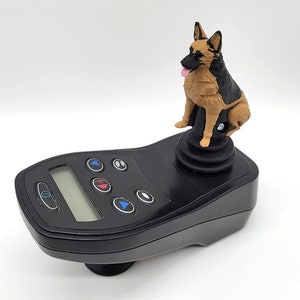 May include: A black joystick with a digital display and several buttons. A brown and black German Shepherd figurine is mounted on top of the joystick.