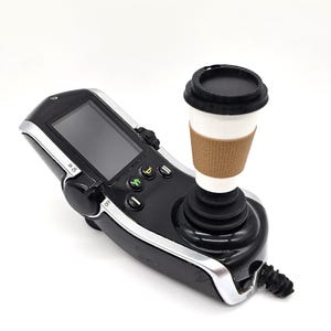 May include: A black gaming joystick with a screen and buttons. A black and white coffee cup sits on top of the joystick.