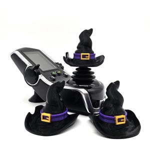 May include: Three black witch hats with purple and gold accents are placed on top of a black joystick controller. The controller has a screen and buttons.