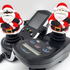 Könnte beinhalten: Zwei Weihnachtsmann-Figuren, eine schwarze und eine weiße, sind an Joysticks befestigt. Die Joysticks sind mit einem schwarzen Gerät mit Bildschirm und Knöpfen verbunden.
