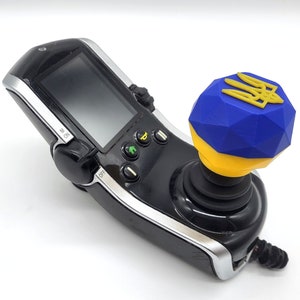 May include: A black and silver mobility device joystick with a blue and yellow geometric knob featuring a gold emblem. The device has a screen and several buttons.