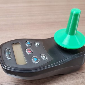 Peut inclure: Une télécommande noire avec un écran numérique et plusieurs boutons. Un joystick vert est fixé au sommet de la télécommande.
