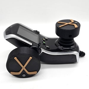 May include: A black and silver joystick controller with a black and gold hockey stick design on the top and bottom. The controller has a screen and buttons.