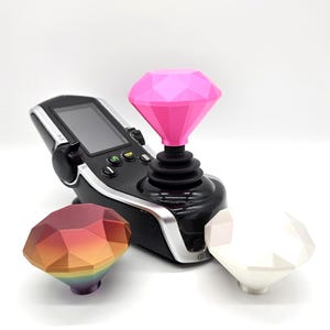 Peut inclure: Une manette de jeu noire avec un dessus en forme de diamant rose. Deux dessus en forme de diamant supplémentaires, arc-en-ciel et blanc, sont également présents. La manette a un écran et des boutons.
