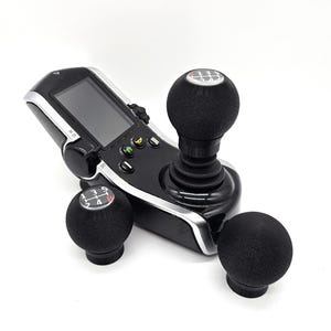 May include: Black gaming controller with a screen, joystick, and three black gear shift knobs. The controller has silver trim and green, yellow, and blue buttons. The gear shift knobs have white markings.