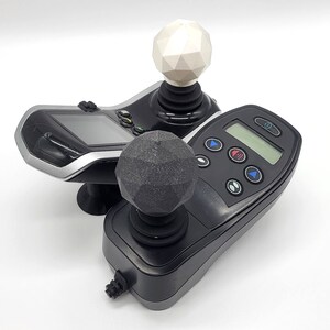 May include: Two black joysticks with different coloured knobs. The top knob is white and the bottom knob is grey. Both joysticks have a black base and a black control panel with buttons.