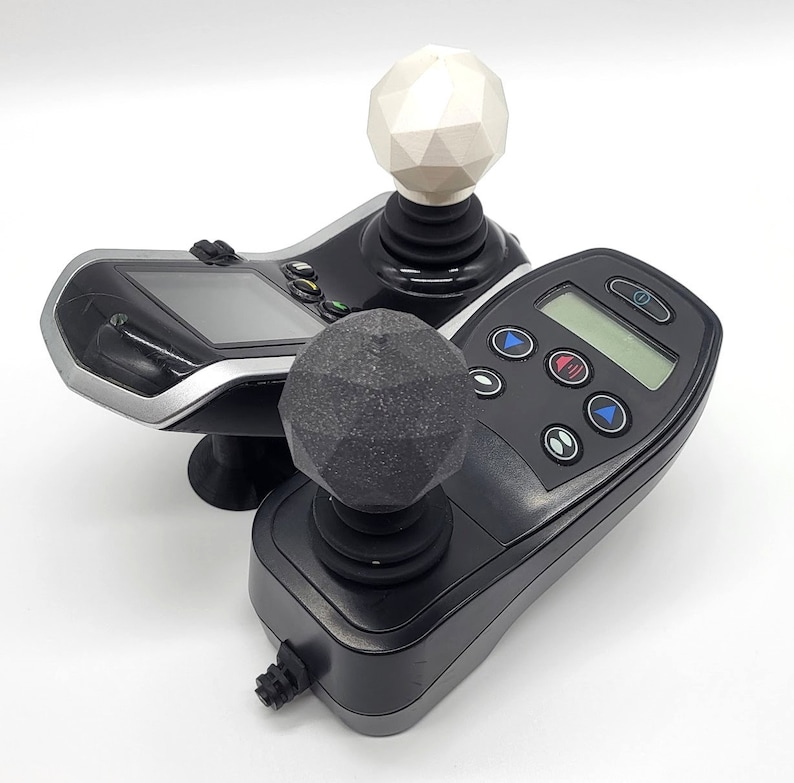 May include: Two black joysticks with different coloured knobs. The top knob is white and the bottom knob is grey. Both joysticks have a black base and a black control panel with buttons.
