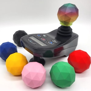 May include: A black electronic joystick controller with a digital display and six buttons, featuring a removable, multi-colored, geometric-faceted topper. Surrounding the controller are six additional solid-colored, faceted spherical toppers in black, blue, yellow, pink, green, and red.