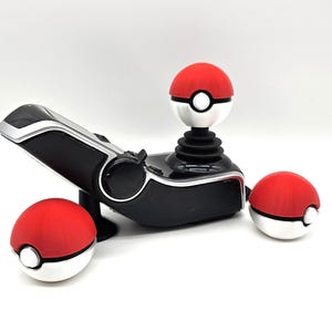 May include: A black gaming controller featuring a joystick topped with a red and white spherical ball, accompanied by two additional red and white spherical balls positioned on either side of the controller base on a plain white background.